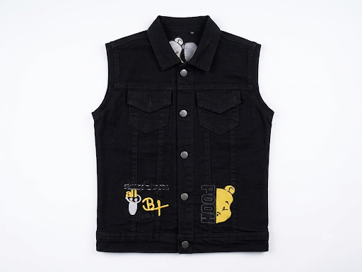 A black denim vest with a button-up front, two chest flap pockets, and graphic patches on the lower front. The left patch features a white and yellow cartoon character with 'B'T' and 'SWEAT' text, and the right patch shows a yellow cartoon bear with 'POO' text.