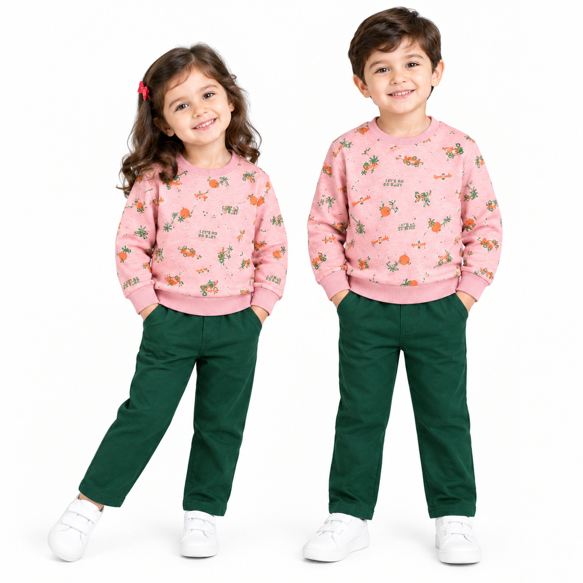 A young girl and boy modeling matching outfits. Both children are wearing pink crewneck sweatshirts with a colorful cartoon animal print, paired with dark green pants and white sneakers, standing against a solid white background.