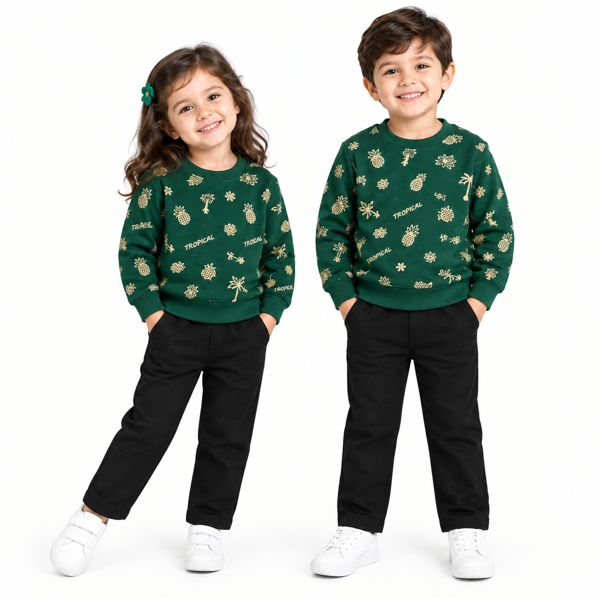 Two children modeling matching dark green crewneck sweatshirts with a gold tropical pineapple pattern, paired with black pants and white sneakers, against a plain white background.